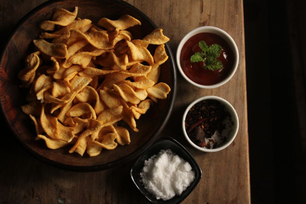 A bowl of crunchy chips paired with spicy and salty dips on a rustic wooden table.
