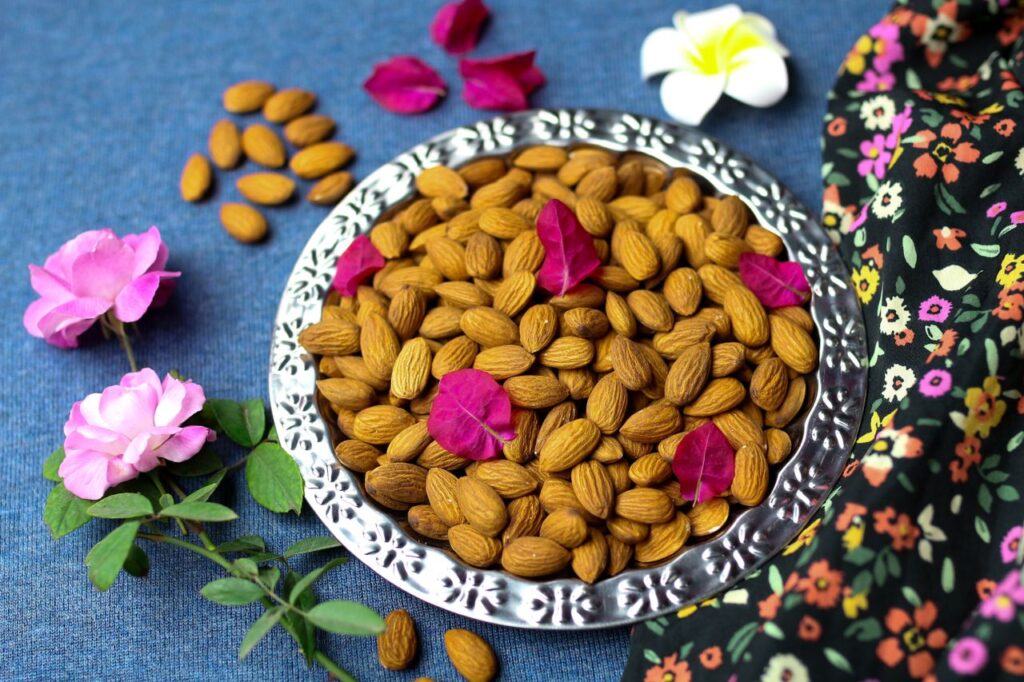 A bowl of almonds adorned with flower petals on a textured surface, surrounded by pink flowers.